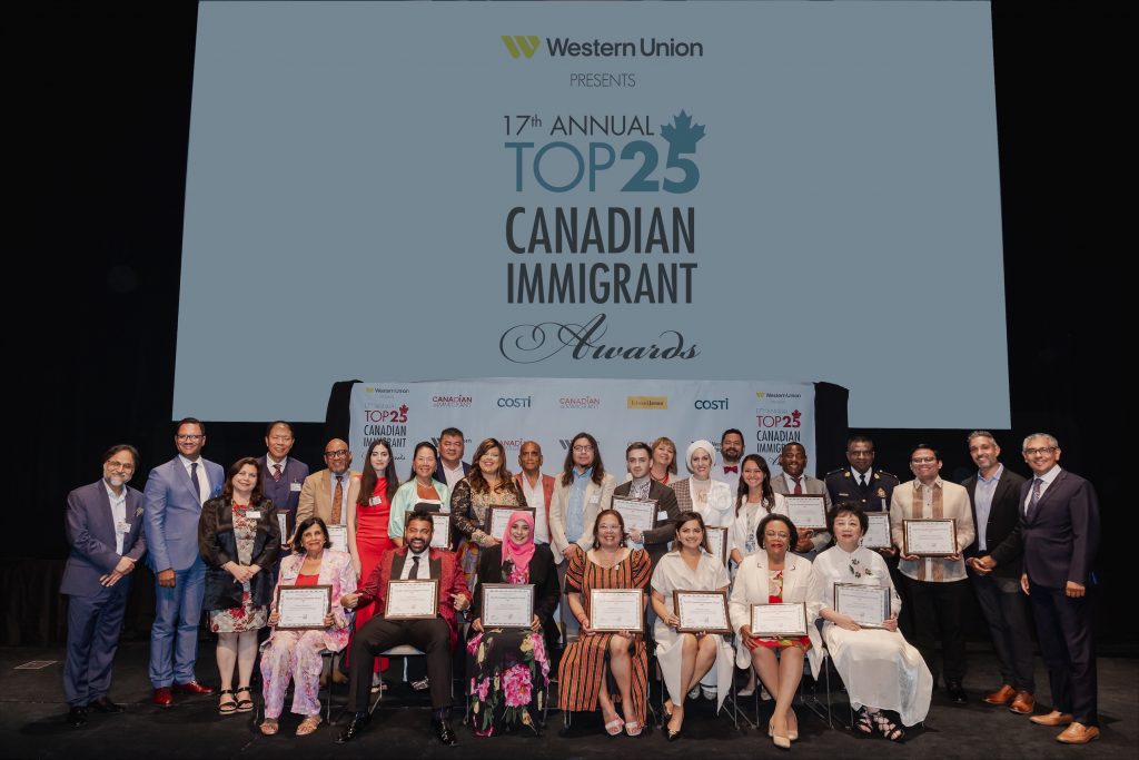 A group of about 25 people on a stage with "Top 25 Immigrant" award logo projected behind them.
