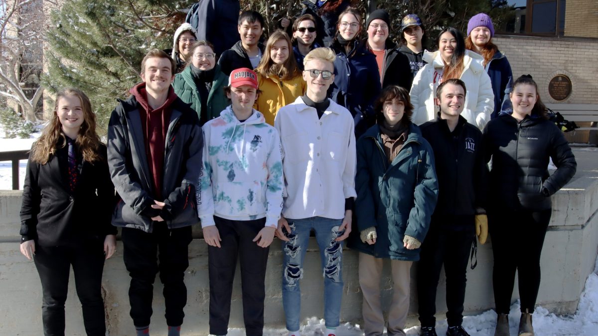A group of students in casual dress stand outside the School of Music building at Brandon University.