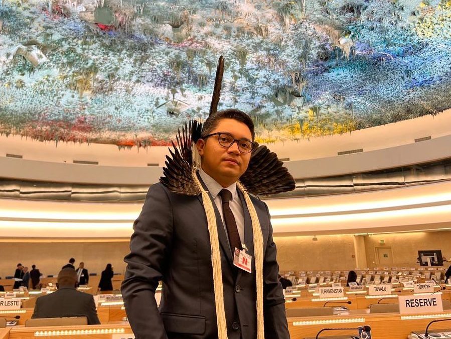A man wearing a traditional headdress stands in a conference room with a colourful ceiling.