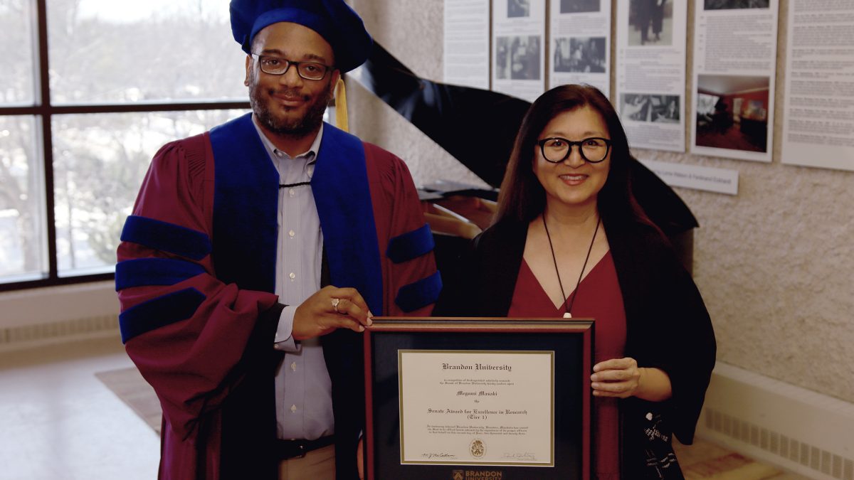 Two people in ceremonial academic robes stand with a framed award certificate.