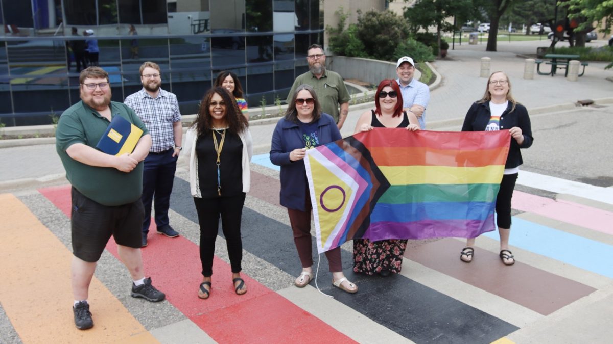 A group of people stand with a large Pride flag on a multi-coloured crosswalk.