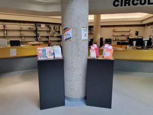 Two tables next to a concrete pillar with books displayed on them. A library circulation desk is in the background.