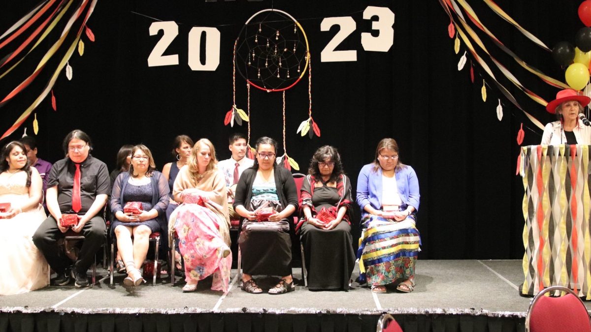 A woman stands and speaks at on a stage. Several people sit to her right.