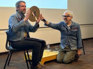 A man is seated and holding a gold cymbal, while a man kneels next to him and holds a microphone. Both men are smiling.