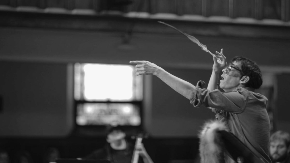 A man points with one hand and holds a feather in his other hand as he conducts musicians