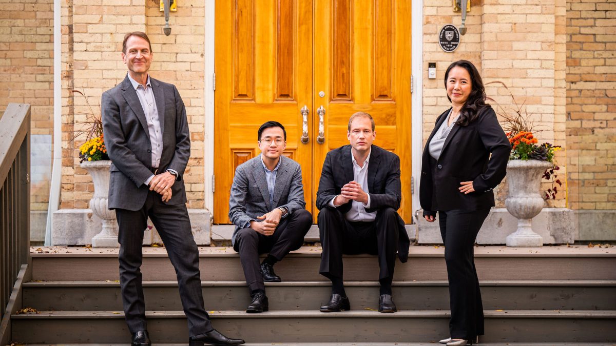 Two men are seated on steps in front of a wooden door. A man stands on their right, while a women stands on their left.