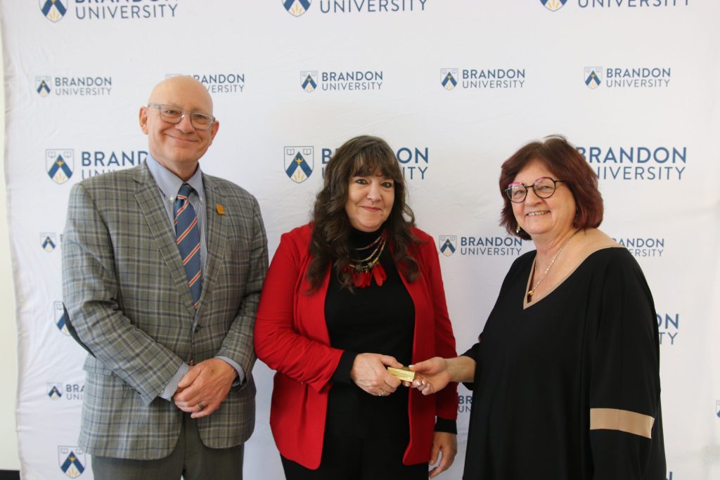 Three people post in front of a backdrop with the Brandon University logo.