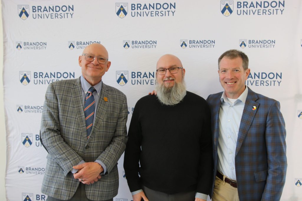 Three people post in front of a backdrop with the Brandon University logo.