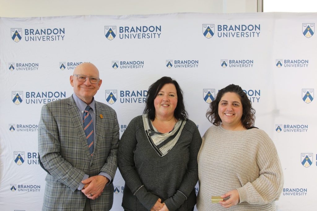Three people post in front of a backdrop with the Brandon University logo.