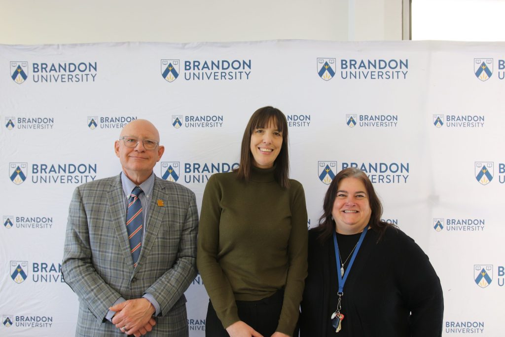 Three people post in front of a backdrop with the Brandon University logo.