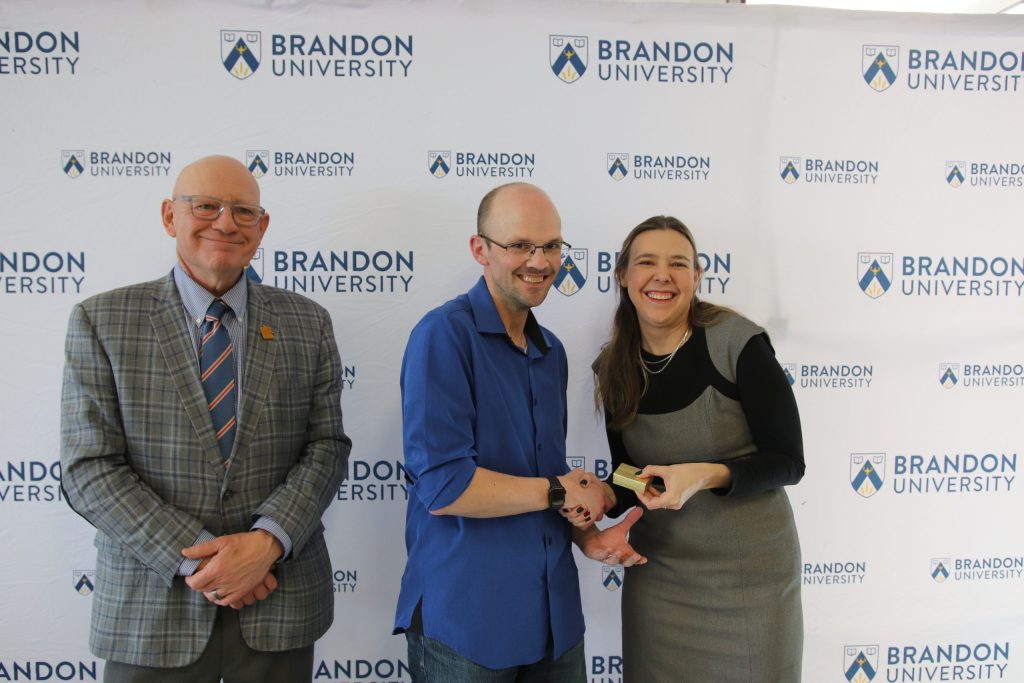 Three people post in front of a backdrop with the Brandon University logo.