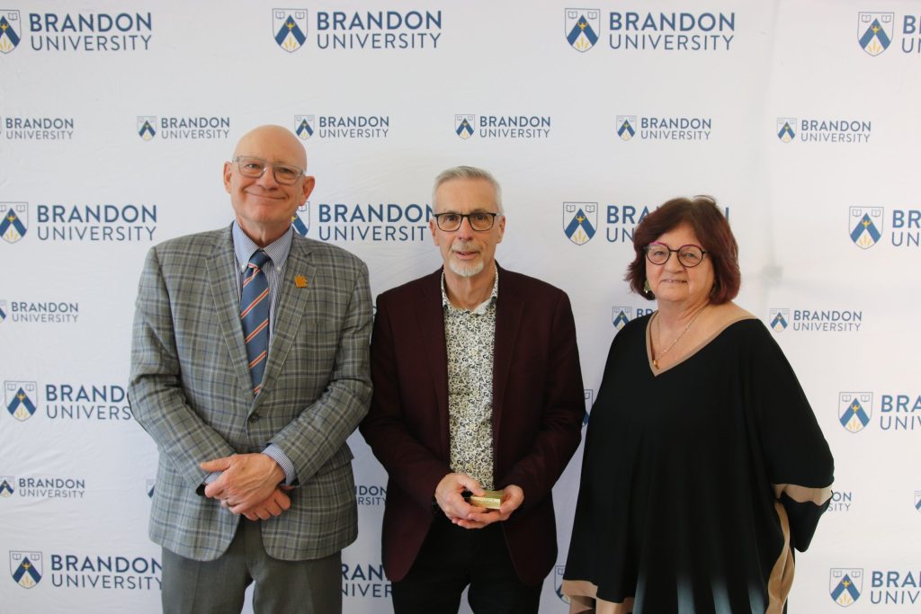 Three people post in front of a backdrop with the Brandon University logo.