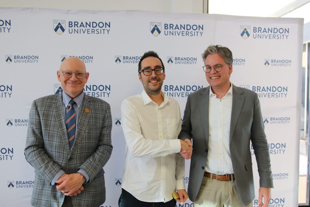 Three people post in front of a backdrop with the Brandon University logo.
