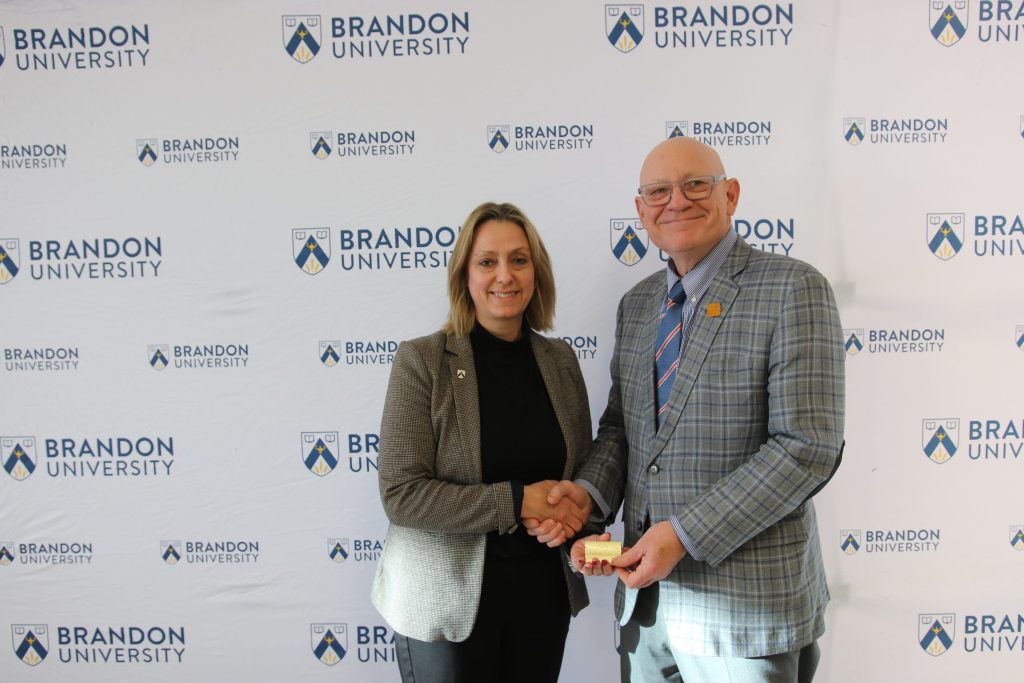 Three people post in front of a backdrop with the Brandon University logo.