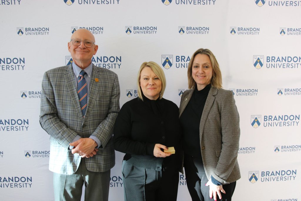 Three people post in front of a backdrop with the Brandon University logo.