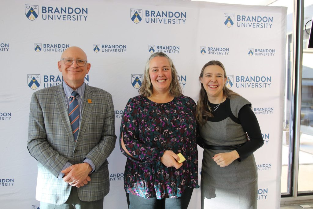 Three people post in front of a backdrop with the Brandon University logo.