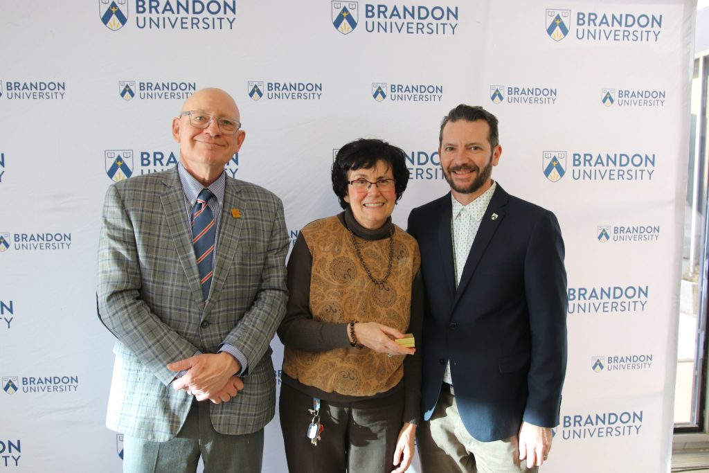 Three people post in front of a backdrop with the Brandon University logo.