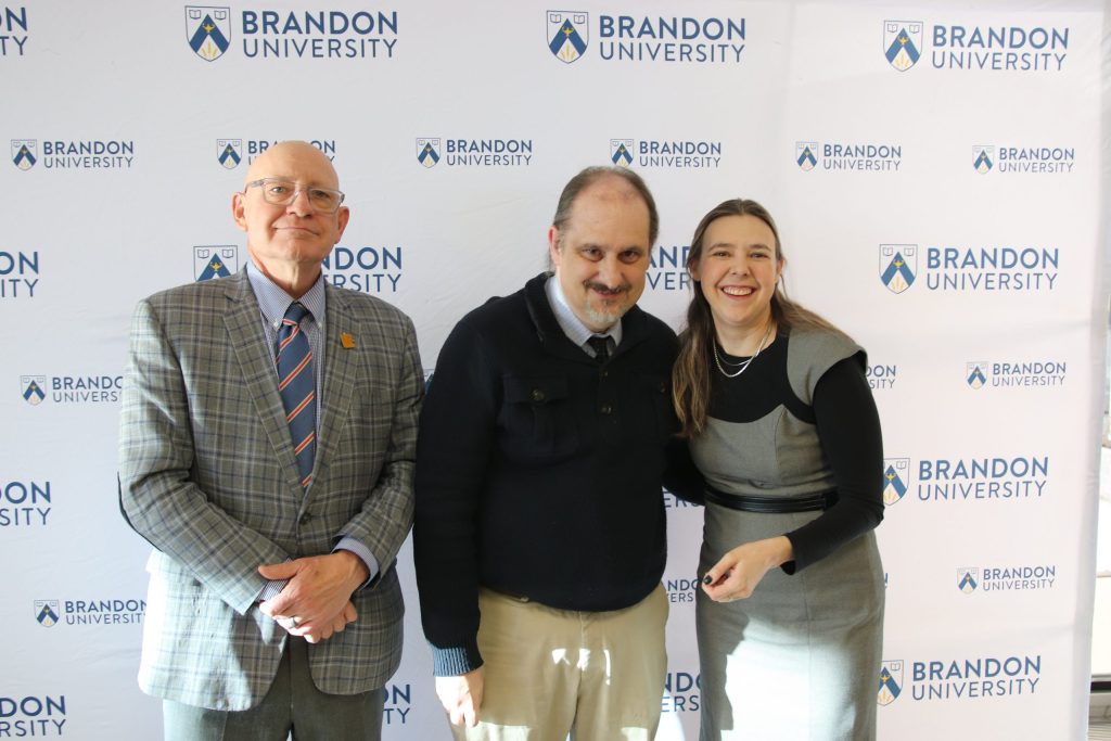 Three people post in front of a backdrop with the Brandon University logo.