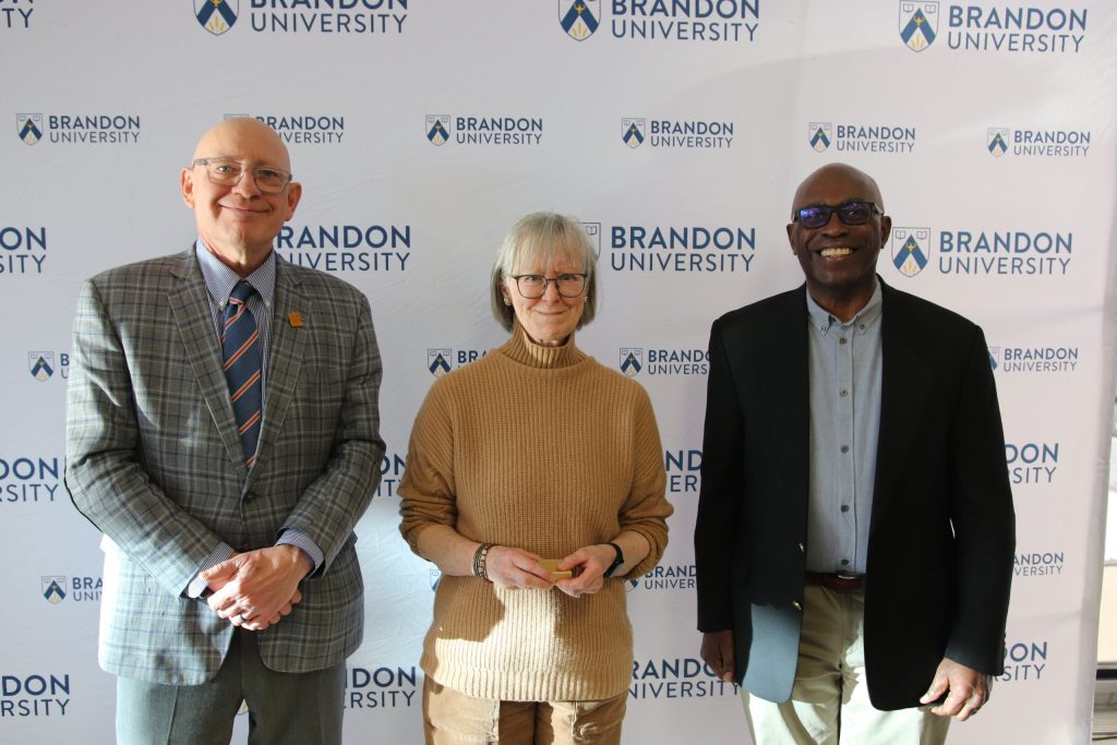Three people post in front of a backdrop with the Brandon University logo.