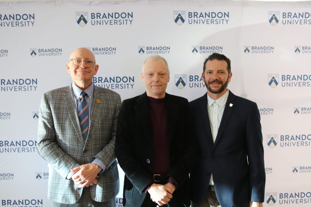 Three people post in front of a backdrop with the Brandon University logo.