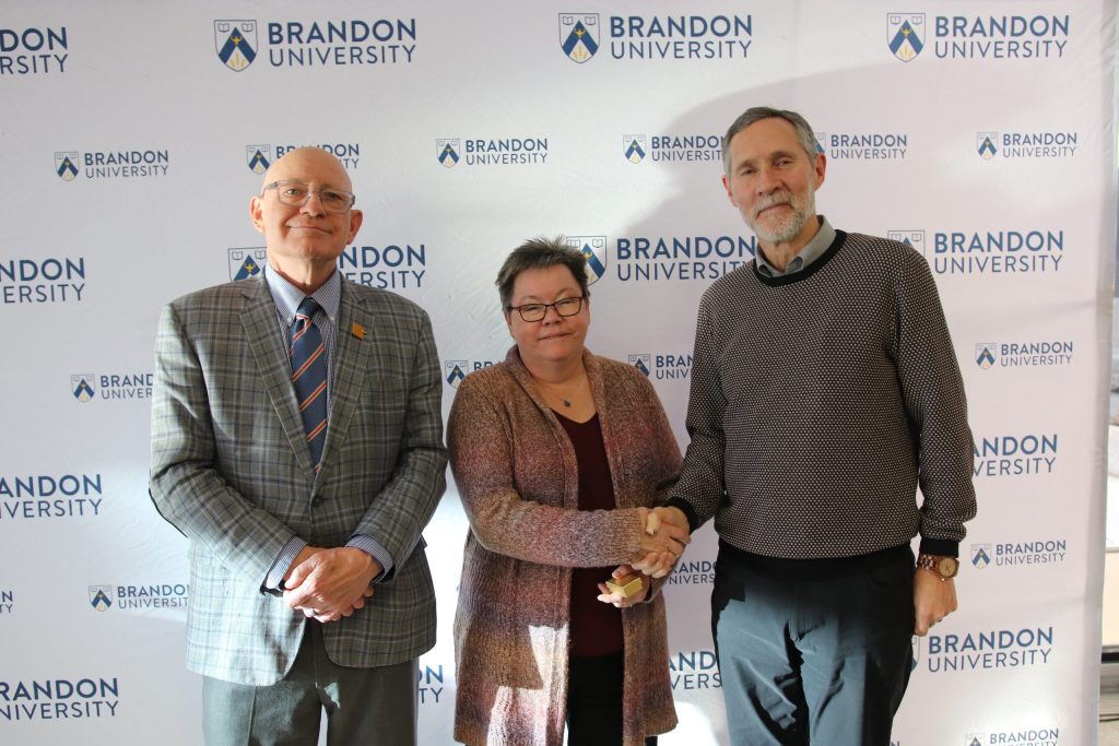 Three people post in front of a backdrop with the Brandon University logo.