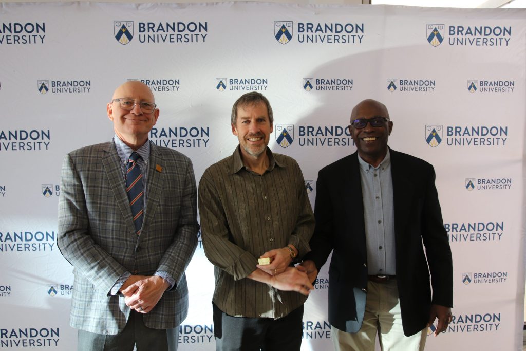 Three people post in front of a backdrop with the Brandon University logo.