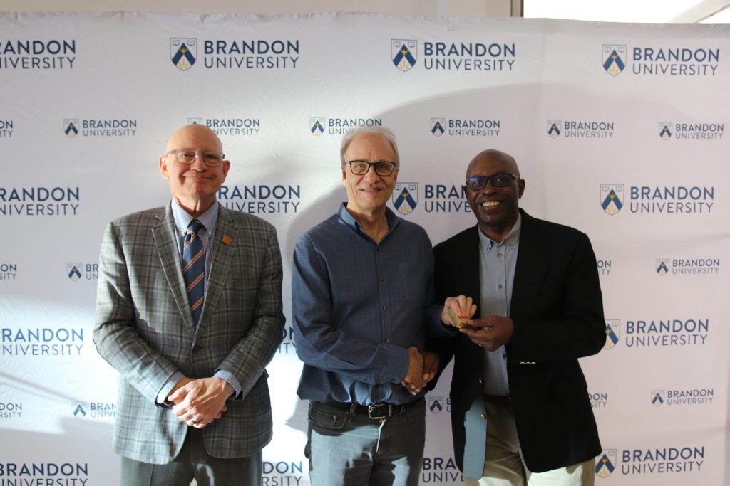 Three people post in front of a backdrop with the Brandon University logo.