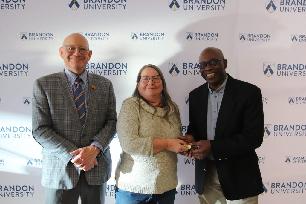 Three people post in front of a backdrop with the Brandon University logo.