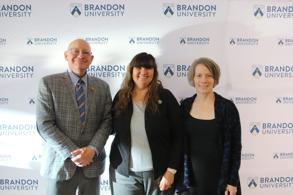 Three people post in front of a backdrop with the Brandon University logo.