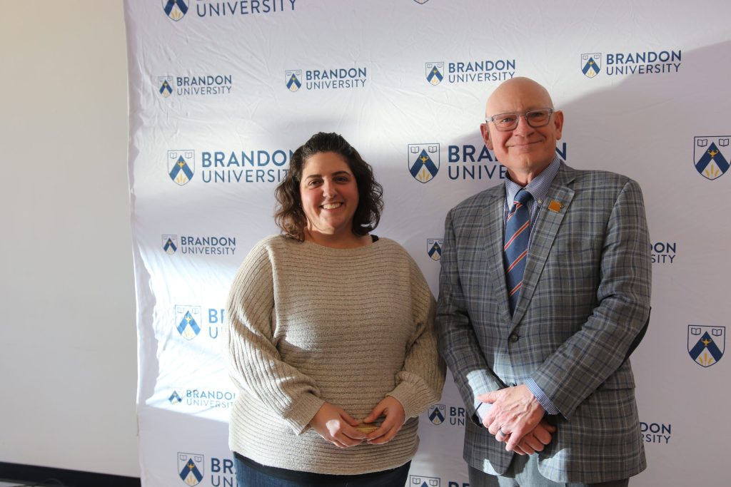 Three people post in front of a backdrop with the Brandon University logo.