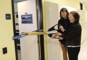 Two women wield oversized scissors to cut a ribbon strung across a door.