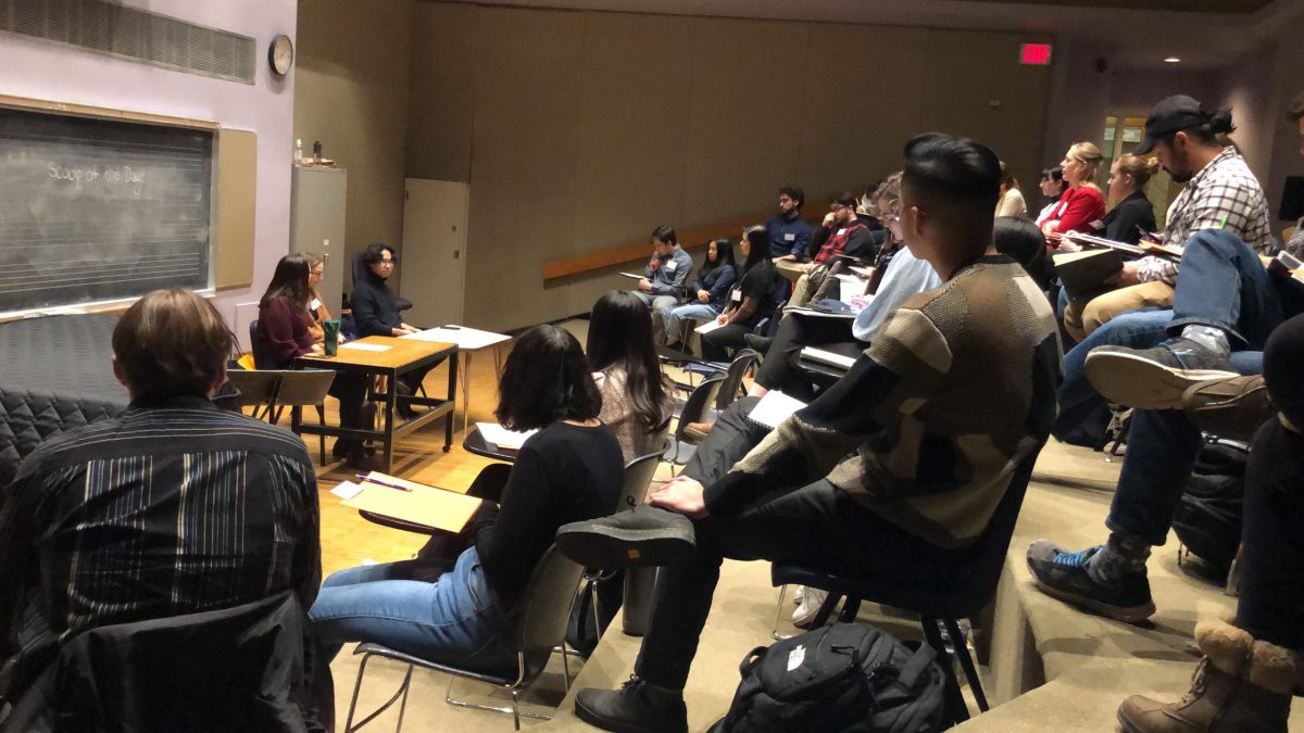 A group of people sit in auditorium-style seating to listen to a presentation from two people at a table in front.