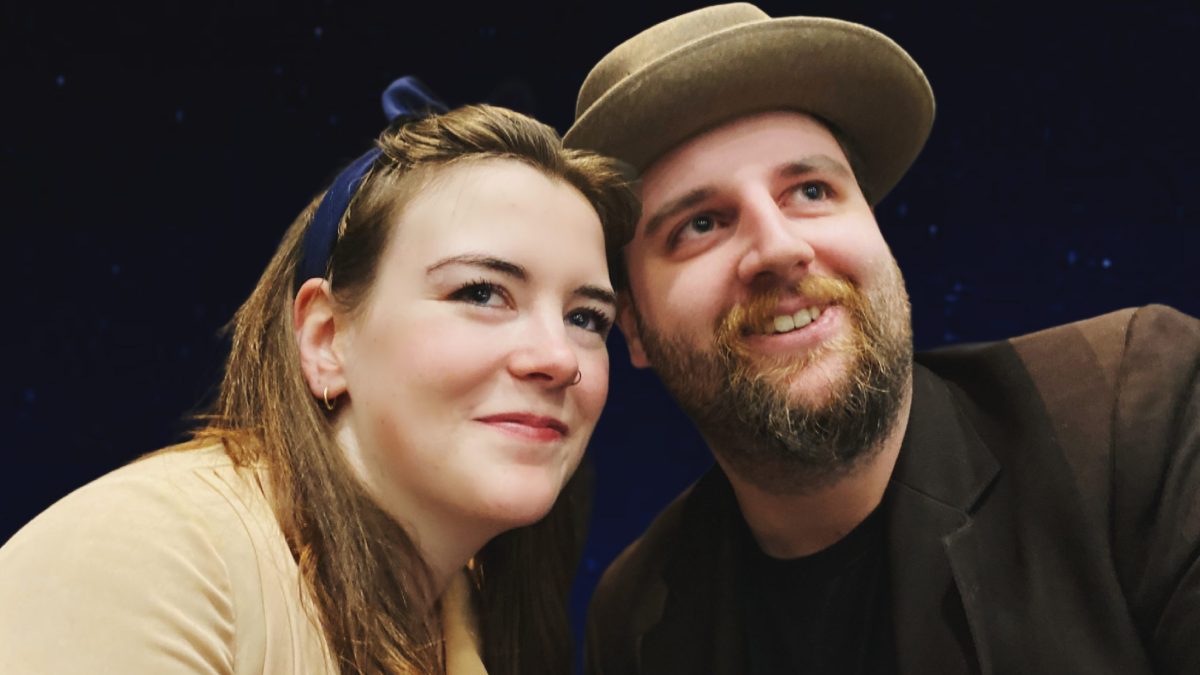 A woman and a man lean close together and smile as they look into the distance