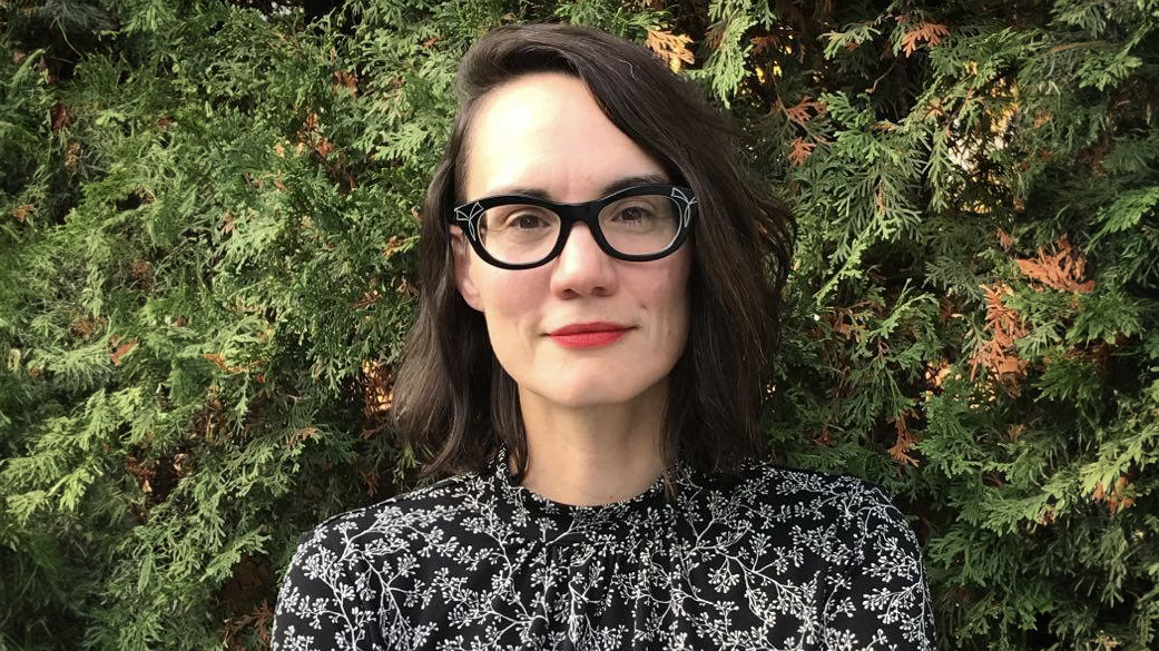 A woman in glasses stands in front of a leafy background.