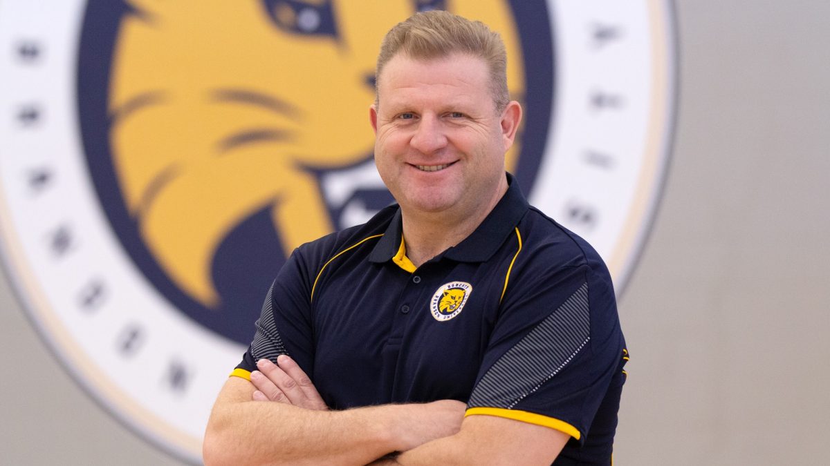 A smiling man poses with his arms folded, with a Bobcats logo on the wall behind him