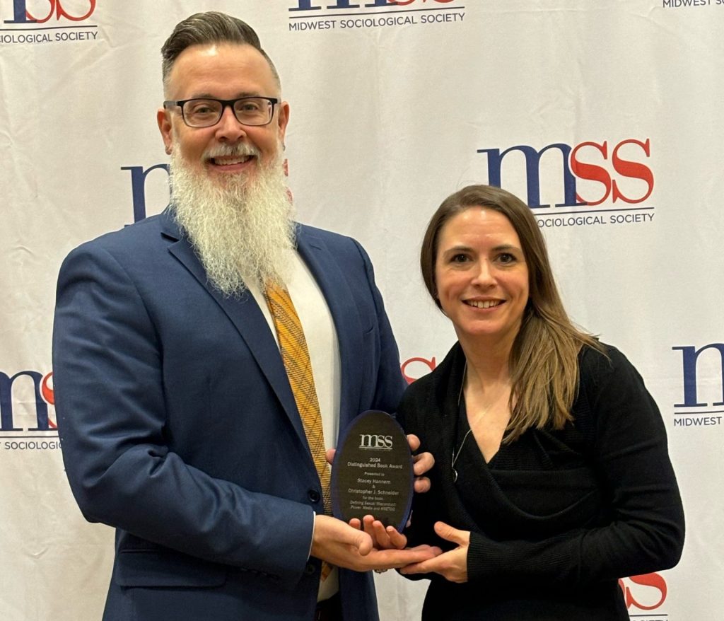A man and a woman smile while holding an award between them