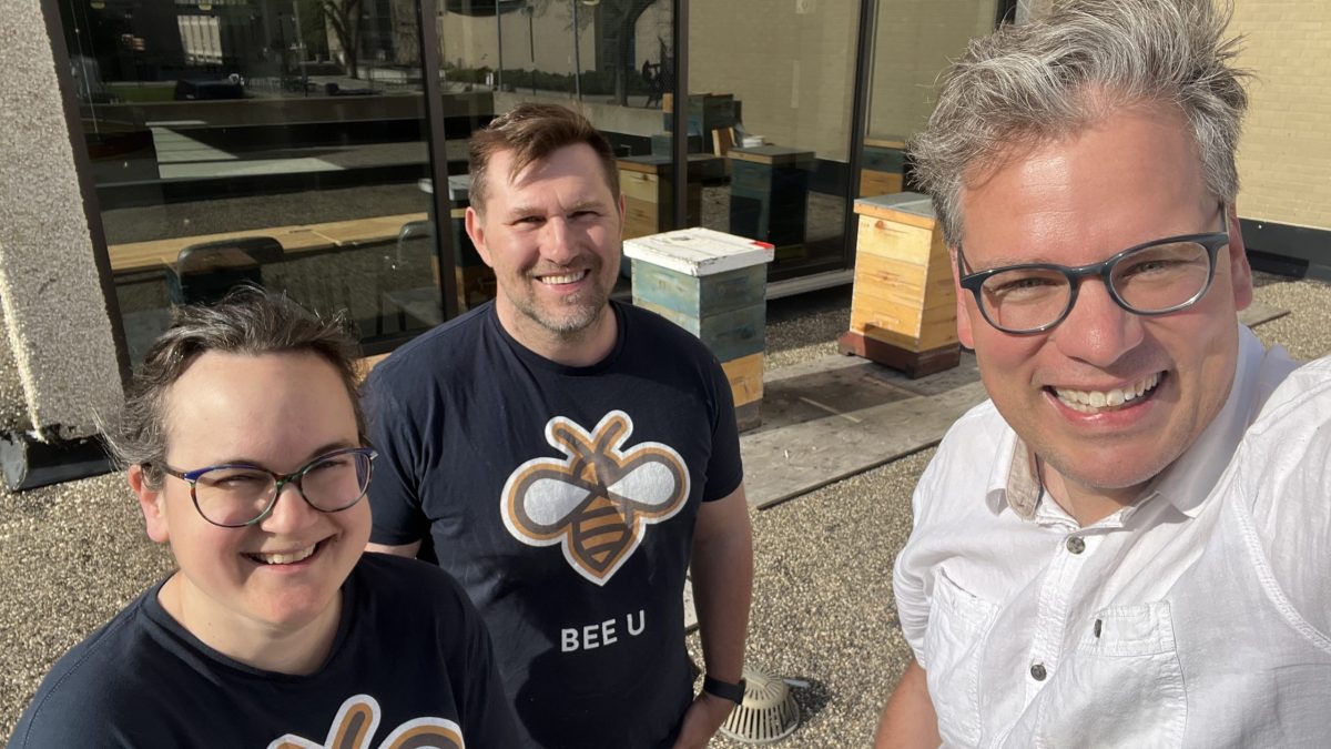 Three people stand on a roof with honeybee hives behind them. Two are wearing "Bee U" shirts.