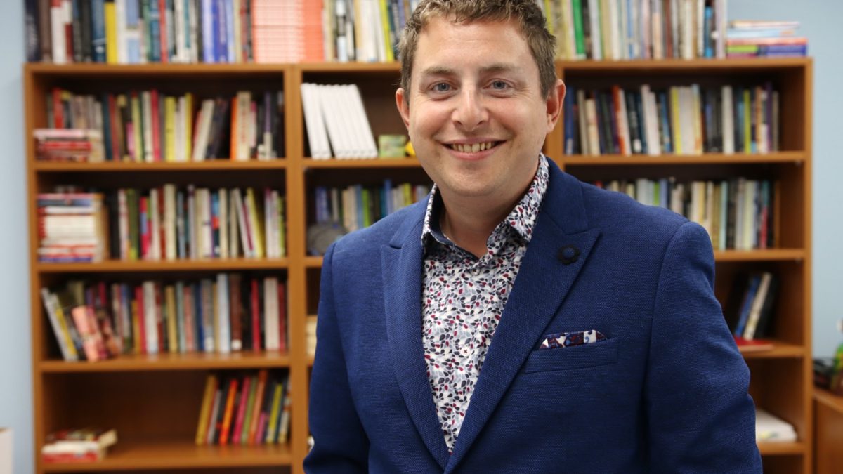 A man in a blue suit stands in front of a full bookshelf.