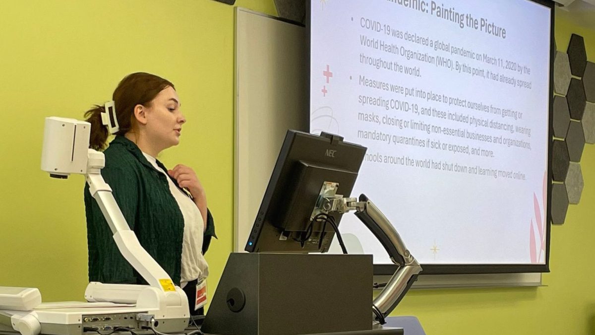 A woman speaks at a podium. Beside her is a projection with the title, "Pandemic: Painting the Picture"