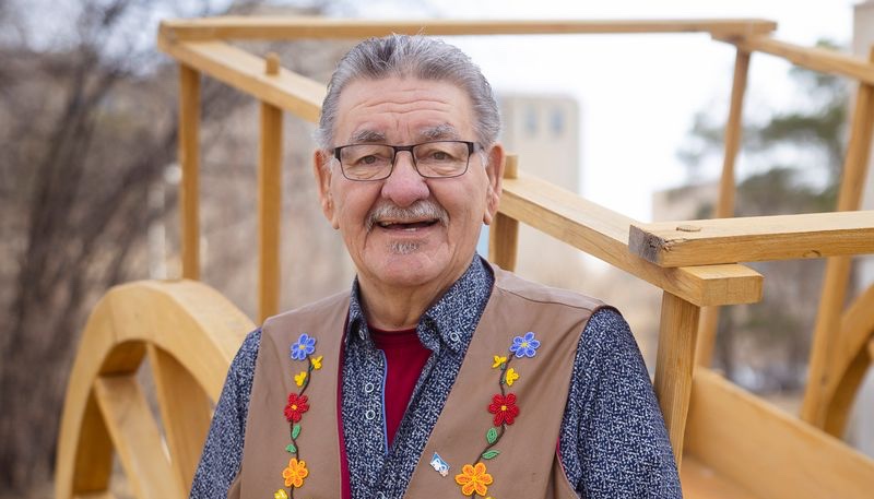 A man in a vest, decorated with beaded flowers, poses in front of a wooden cart