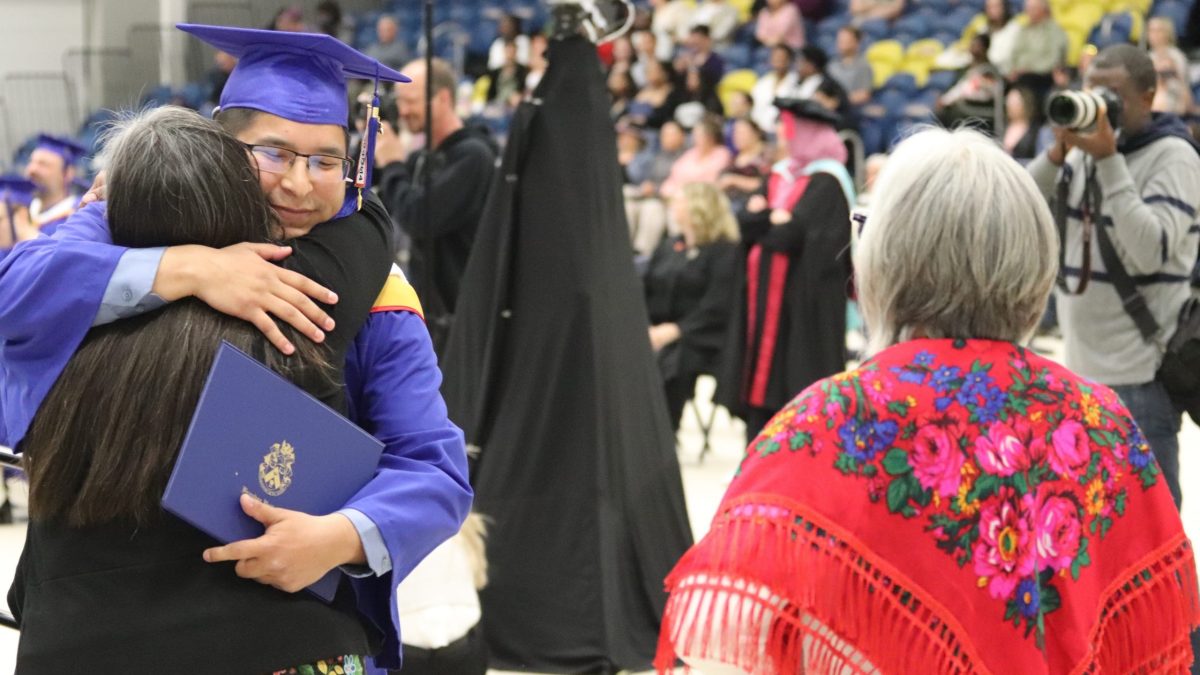 A man in a convocation robe and cap hugs a woman, while another woman looks on