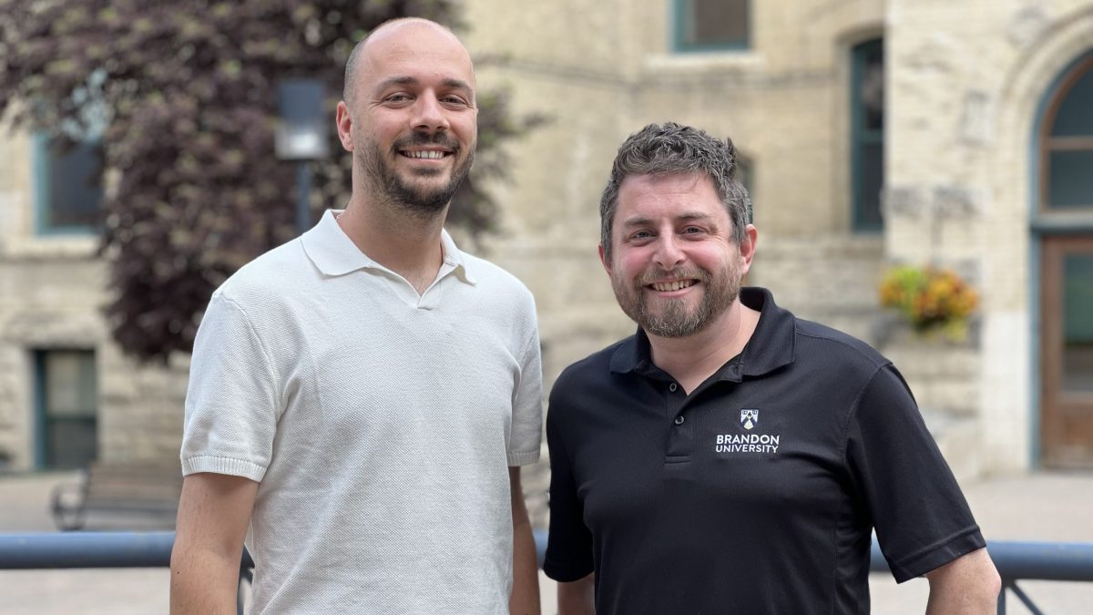 Two men stand in the courtyard of Brandon University. The one one the right wears a BU shirt.