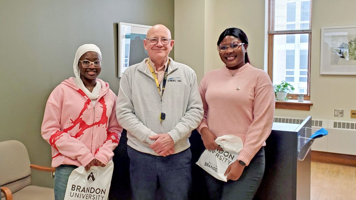 Two women, stand with a man between them. The women are holding Brandon University bags