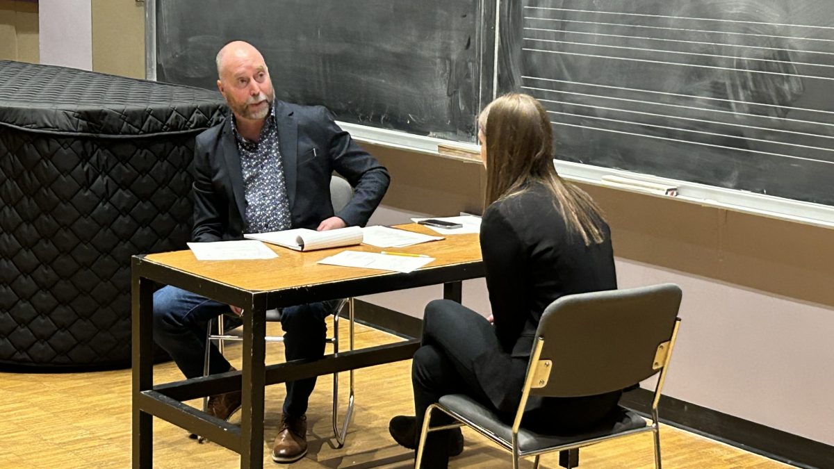 Two people confer at a desk in front of a chalkboard with music notation on it.