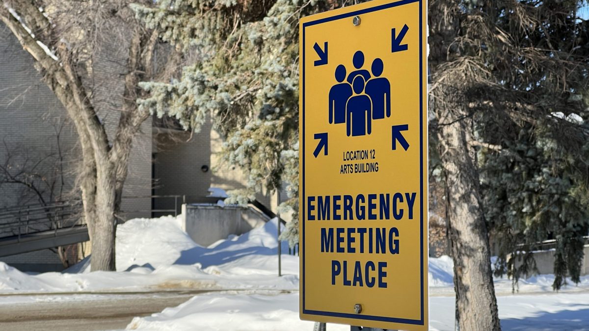 A sign outside of a campus building says "Emergency meeting place" with arrows pointing to a group of people.