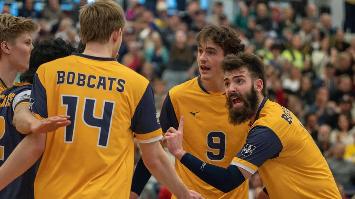 A man with a beard shouts and holds up his index finger as he is surrounded by teammates