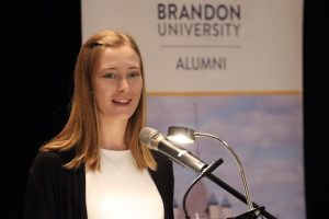 A young woman speaks at a microphone. A banner behind her says "Brandon University Alumni"