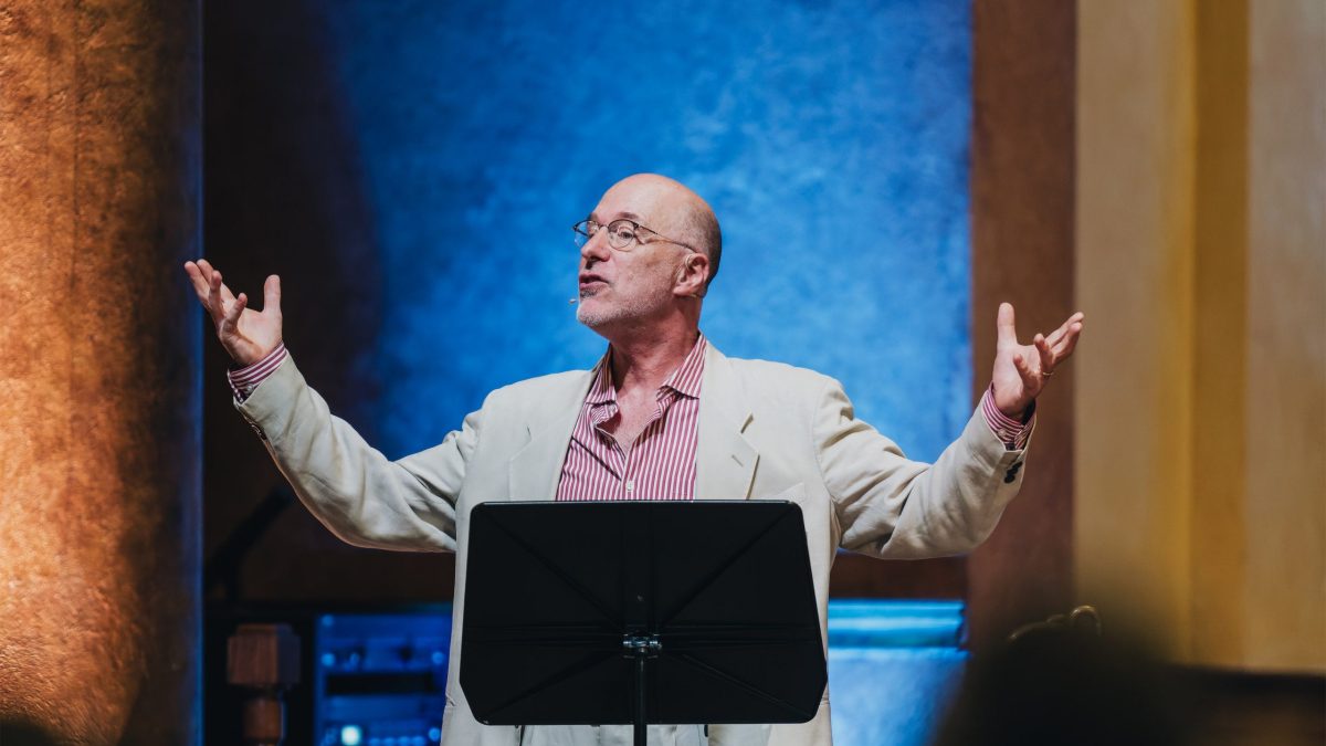 A man raises his arms and looks up while standing behind a music stand