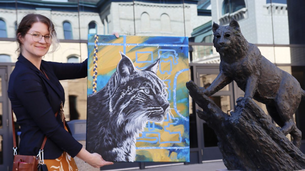 A woman holds up a painting of a bobcat next to a bobcat statue