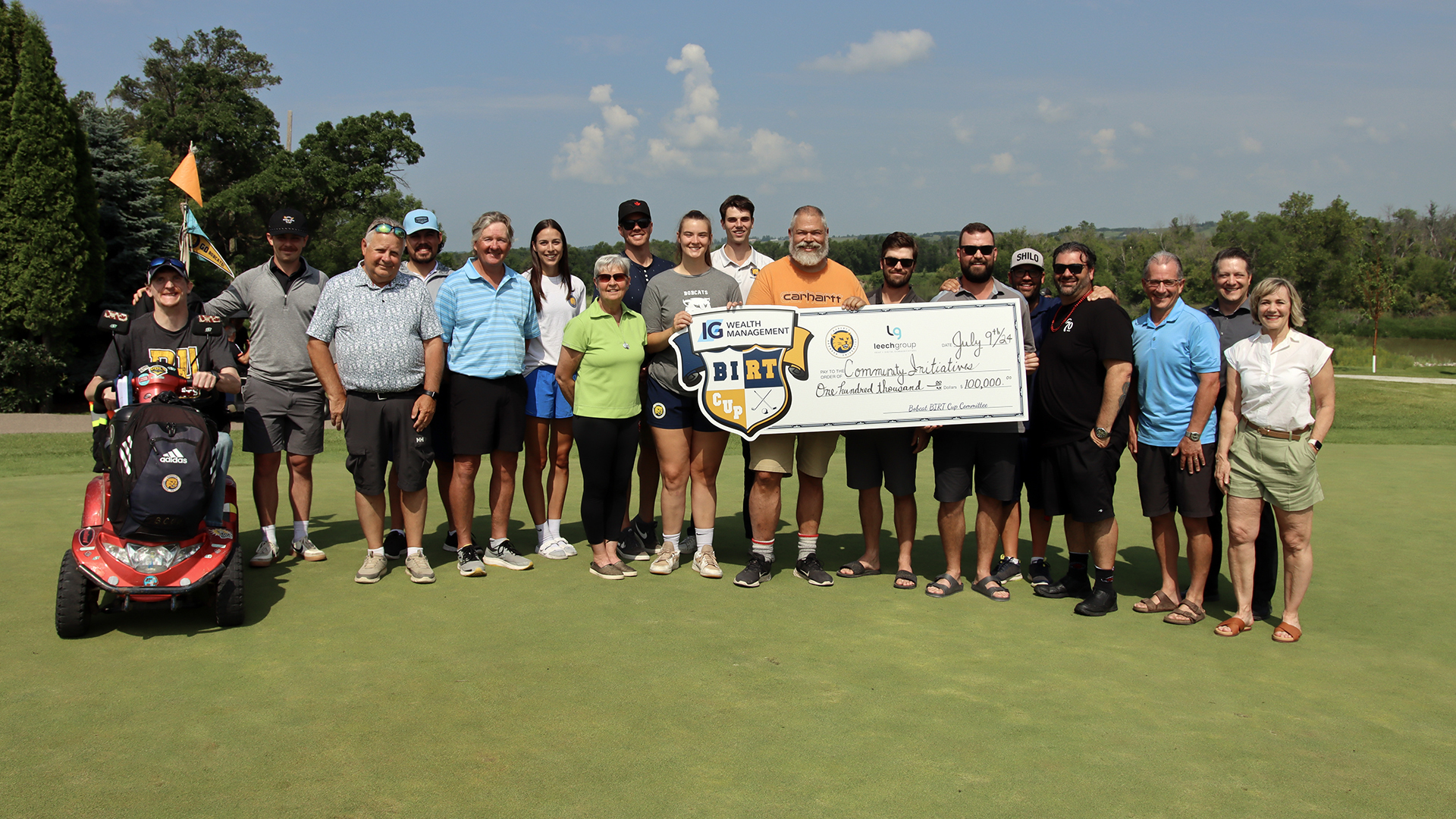 A large group of people stand on a golf tee box, holding an oversized novelty cheque that shows $100,000.